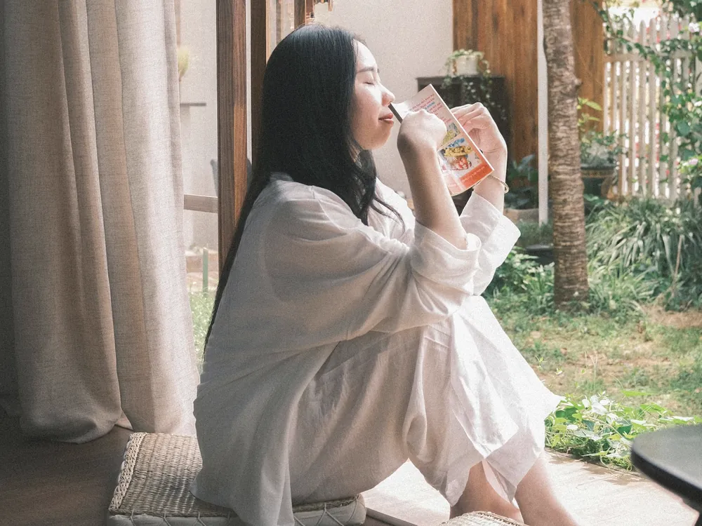 開いた窓のそばで、白い服の女性が床に座ってお茶を飲んでいる