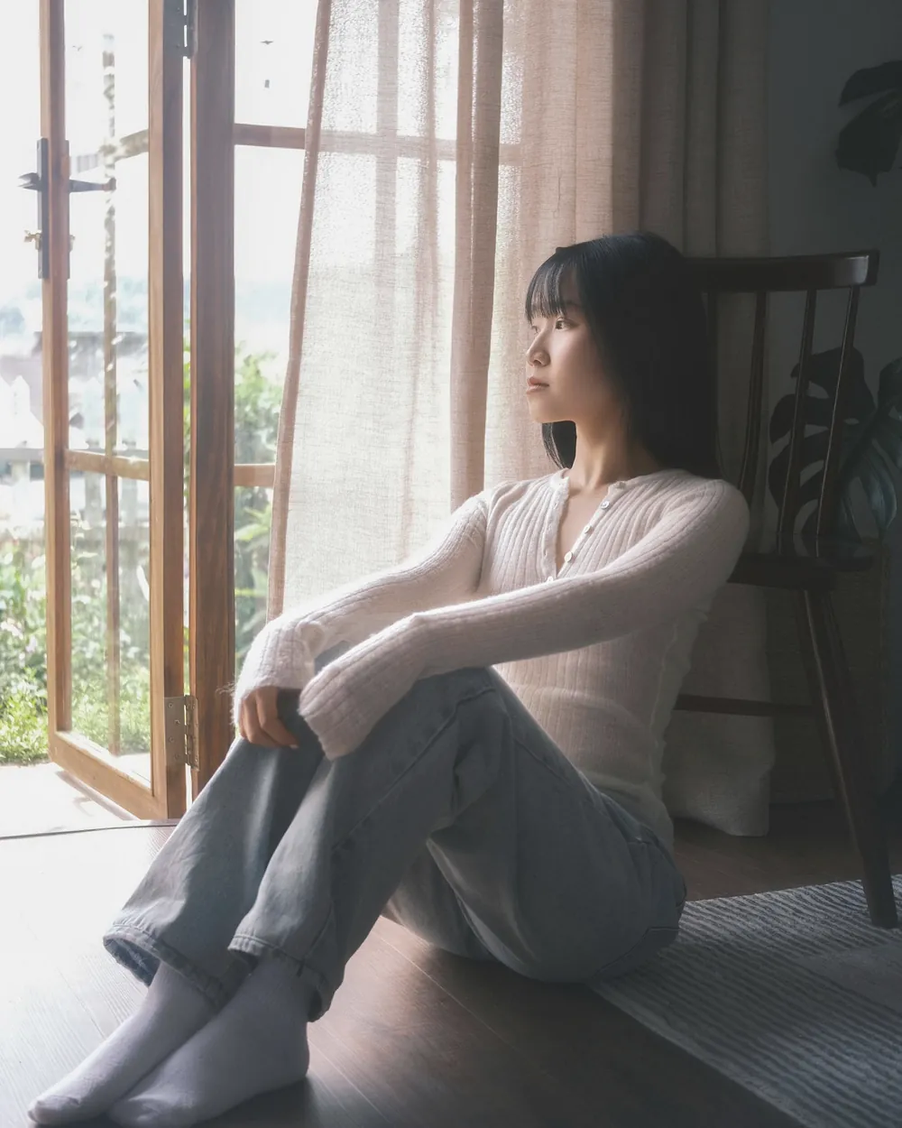 窓のそばの床に座り、膝を抱えて外を見る女性